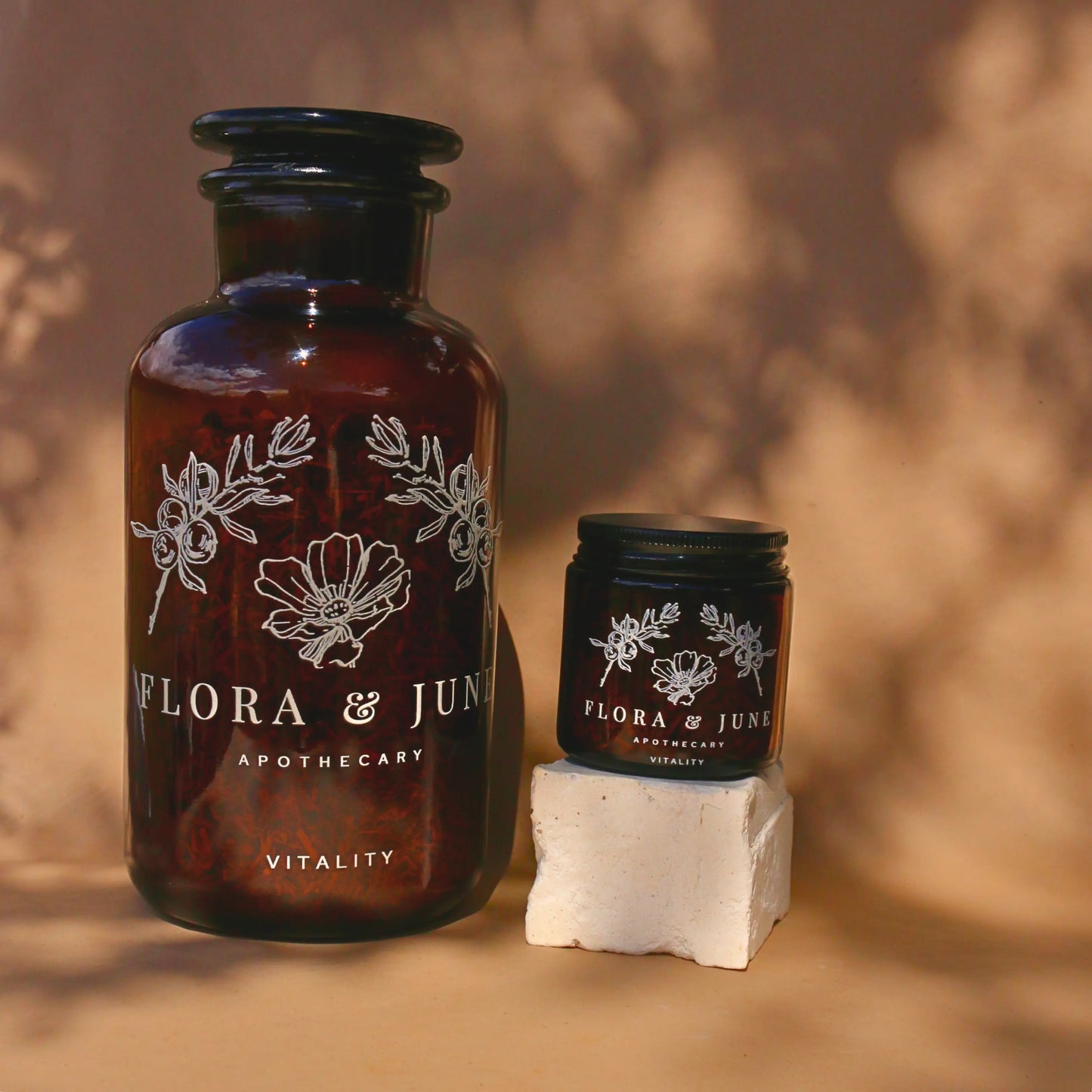 Two sizes of amber glass jars filled with Flora & June's Vitality loose leaf tea. Jars have a white label showing the brand name "Flora & June Apothecary" and the brand logo of a blooming flower and two sprigs of Juniper branch.