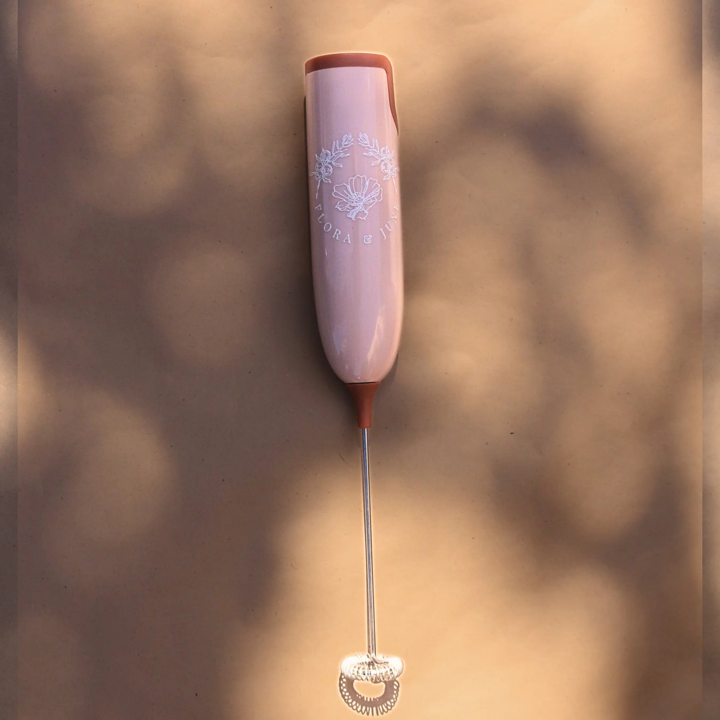 Flora & June electric milk whisk in a soft pink tone with terracotta accents at either end of the handle. Displays the Flora & June logo in white, the the brand name and two juniper branches arching over a blossoming flower in the centre.
