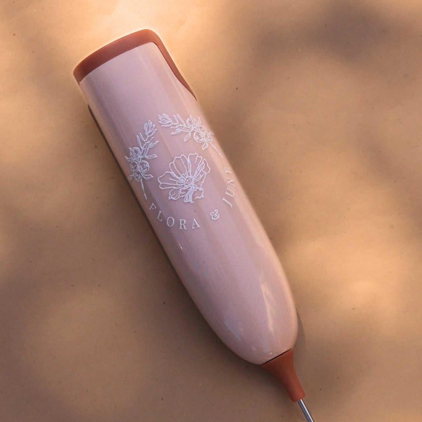 A close up of detail on Flora & June electric milk whisk in a soft pink tone with terracotta accents at either end of the handle. Displays the Flora & June logo in white, the the brand name and two juniper branches arching over a blossoming flower in the centre.