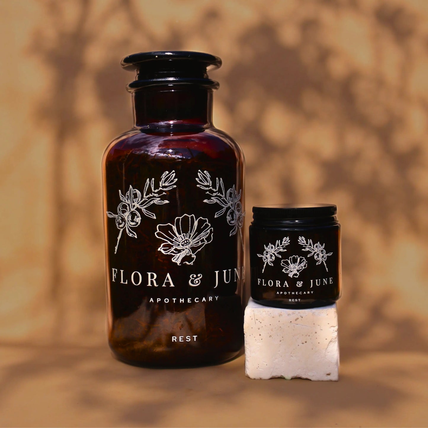 Two sizes of amber glass jars containing Flora & June's 'Rest' loose leaf tea. Larger size in reagent 1000ml apothecary bottle, the smaller size in 100ml jar with black screw top lid. both jars show a white label the the brand name "Flora & June Apothecary", the tea name "Rest", and the brand logo of a blossoming flower and two Juniper branches arching over the flower.