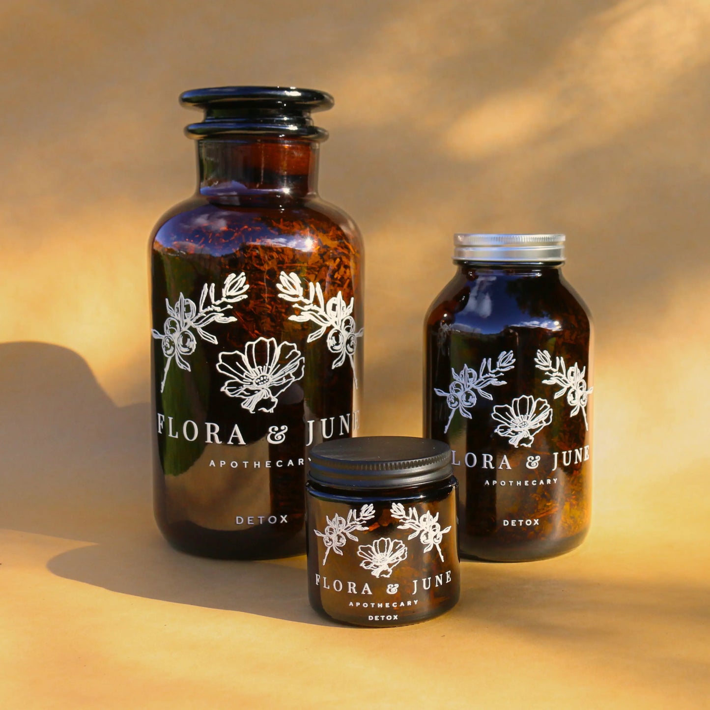 Flora & June's Detox tea in three sizes, with a large amber bottles, set against a backdrop of soft, dappled light.