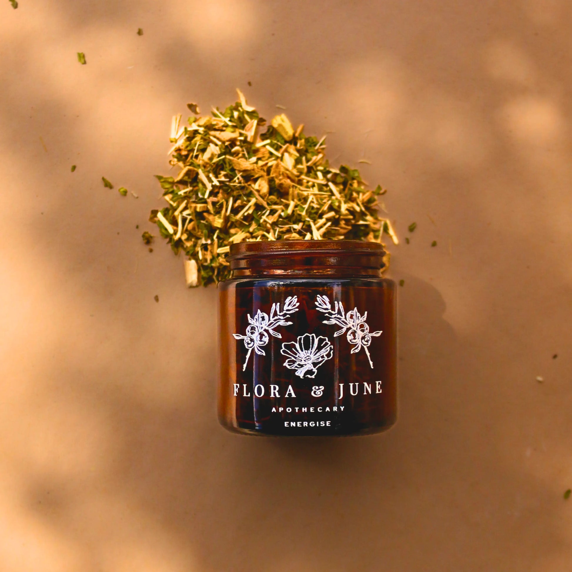 Overhead view of Flora & June's 'Energise' loose leaf tea, spilling vibrantly from an amber jar onto a surface kissed by sunlight and shadows, reflecting the blend's refreshing and invigorating qualities.