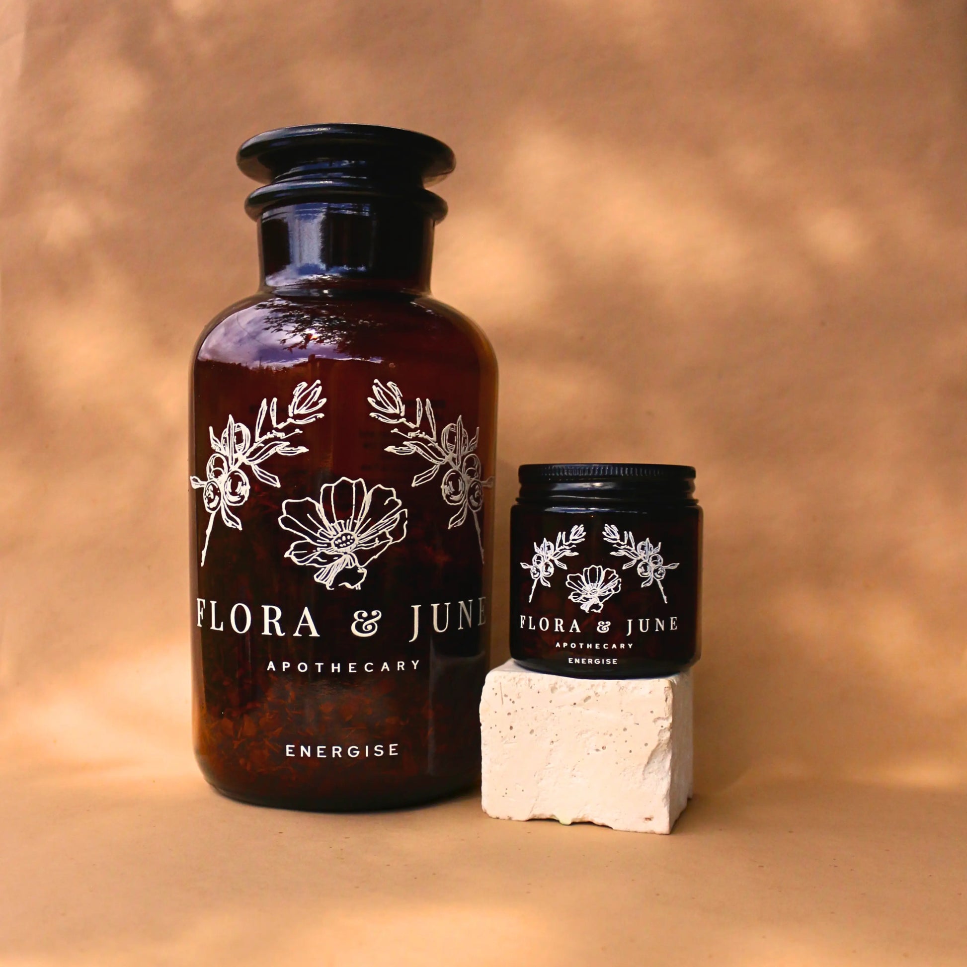 Flora & June Apothecary's 'Energise' blend presented in an amber glass bottle and small black jar, set upon a natural backdrop with gentle shadows, portraying the tea's potential for natural energy enhancement.