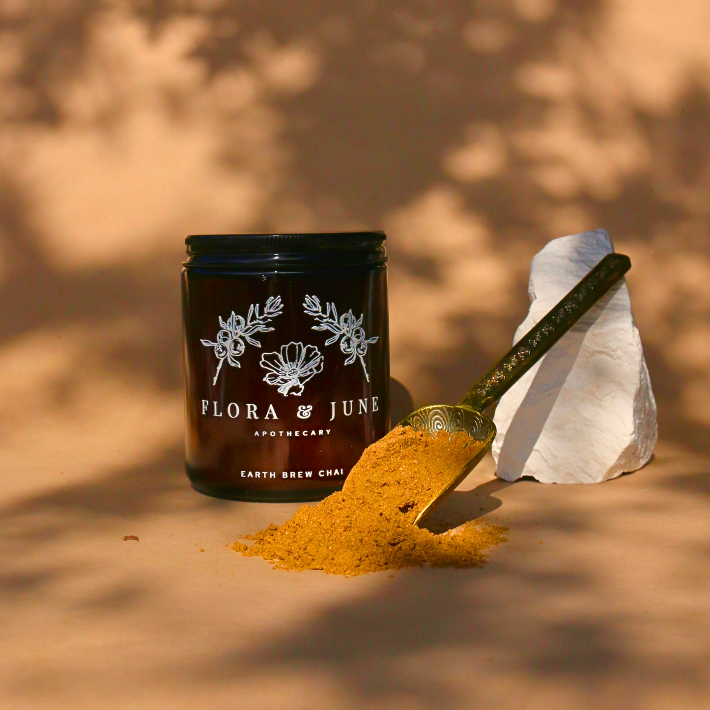 A golden spoon dusted with Flora & June's Earth Brew Chai powder leans against a white stone, with the chai's warm golden hues spilling onto the shadow-speckled surface beside the labeled jar, suggesting a rich, spice-infused morning ritual.