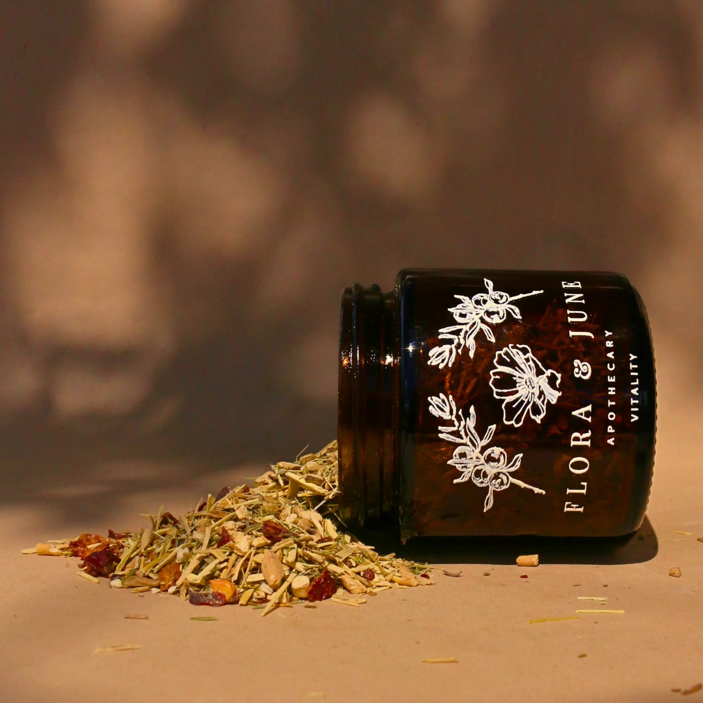 An amber glass jar on it's side with Flora & June's Vitality loose leaf tea spilling out onto the page. The jar has a white label showing the brand name "Flora & June Apothecary" and the brand logo of a blooming flower and two sprigs of Juniper branch.