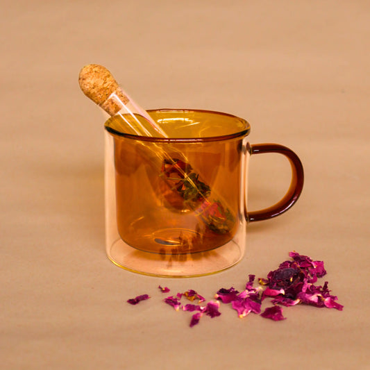 Your perfect tea time companion: our glass infuser with cork, to lock in essential oils released in steam. The infuser, filled with Flora & June's floral 'Balance' blend, sits in an amber glass mug. A scattering of vibrant dried rose petals lay in the foreground.