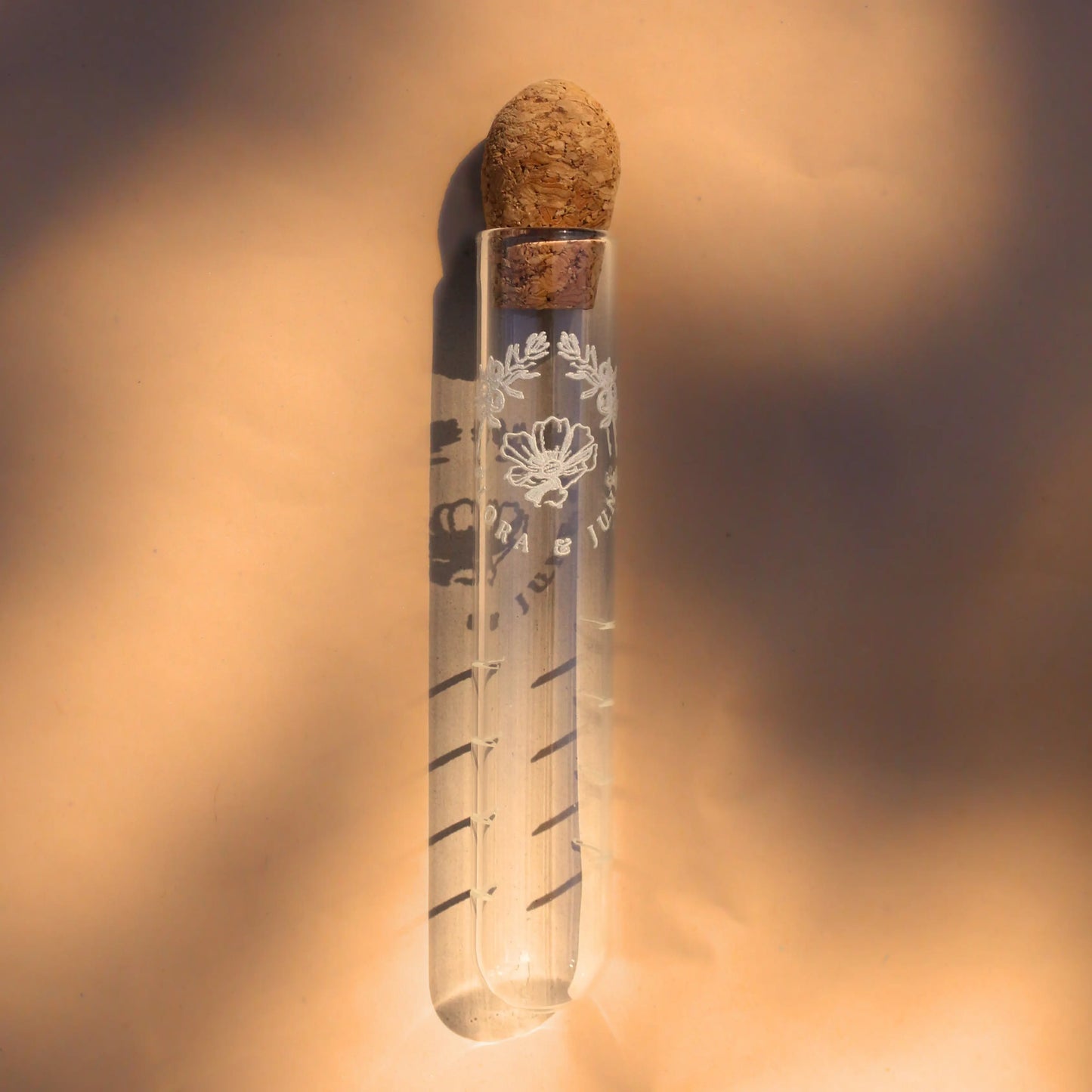 Glass tube infuser displayed in dappled morning sunlight. The infuser is topped with a cork and a white label depicting the brand name "Flora & June" and the logo of two juniper branches arching over a blossoming flower in the centre.