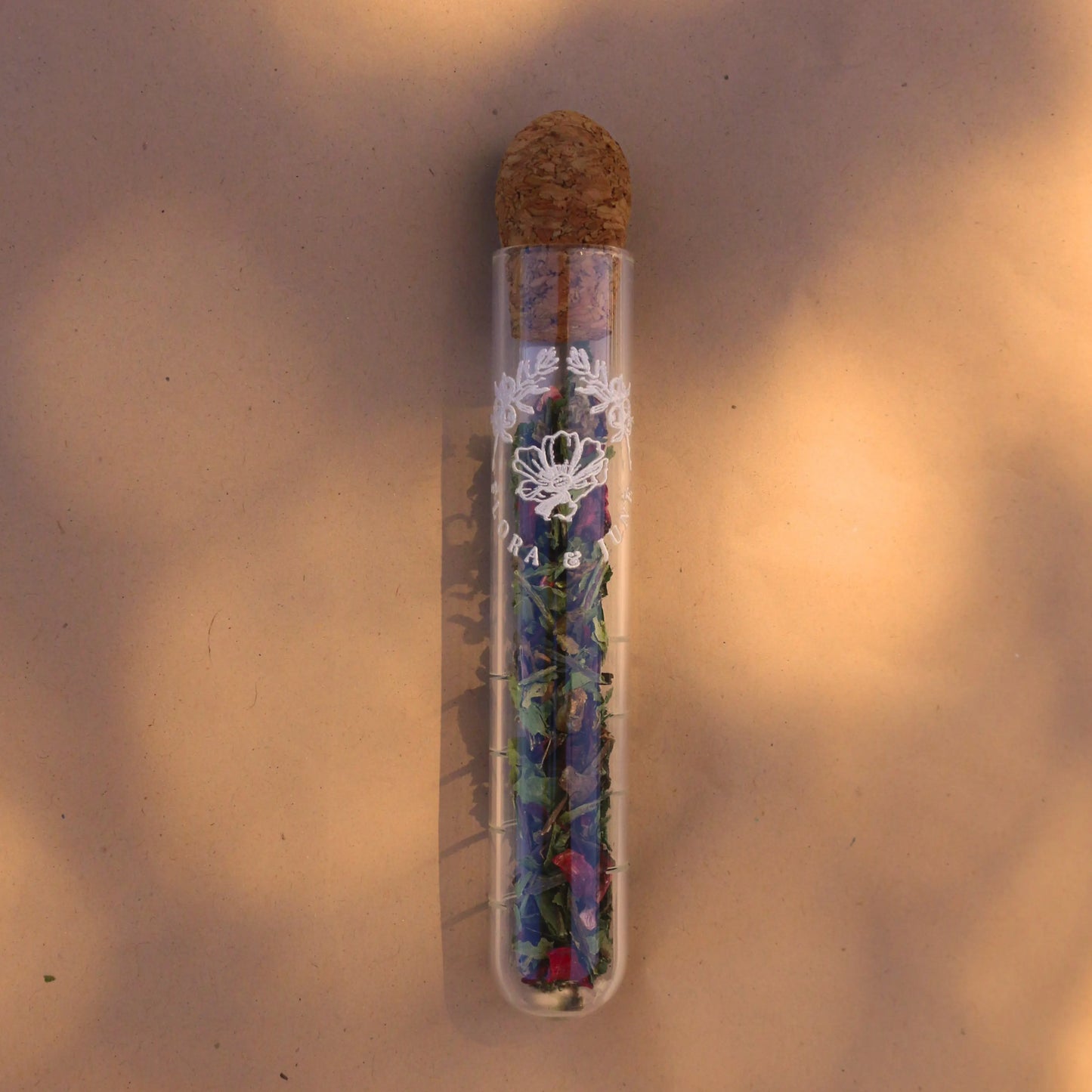 Glass tube infuser displayed in dappled morning sunlight. The infuser is topped with a cork and a white label depicting the brand name "Flora & June" and the logo of two juniper branches arching over a blossoming flower in the centre. The infuser is filled with a sample of Detox loose leaf tea blend.