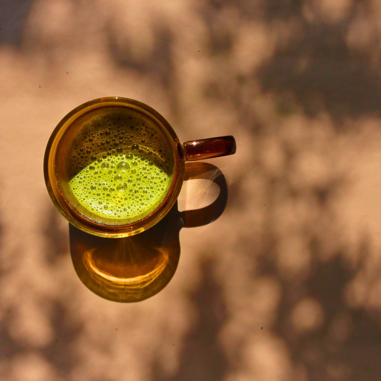 Bathed in dappled shadows, an amber glass mug is filled with Flora & June's vibrant green Matcha Latte 