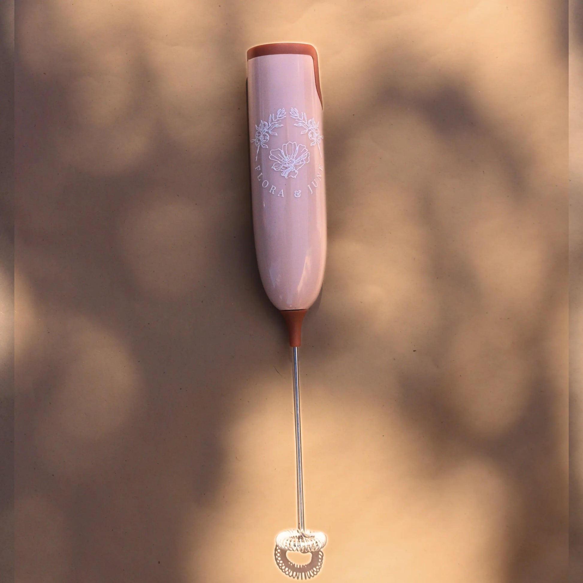 Flora & June electric milk whisk in a soft pink tone with terracotta accents at either end of the handle. Displays the Flora & June logo in white, the the brand name and two juniper branches arching over a blossoming flower in the centre. 