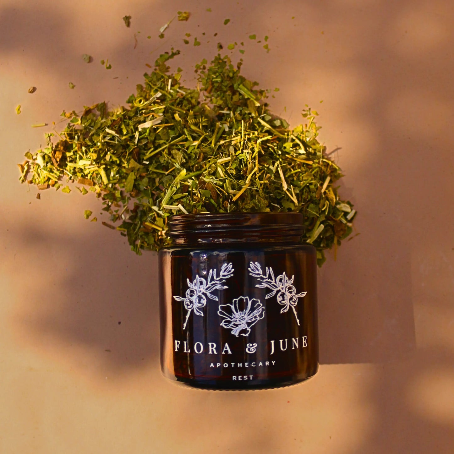 100ml amber glass jar turned on it's side, with Flora & June's Rest loose leaf tea spilling out onto the page. The dappled light is shimmering over the vibrant herbal tea.