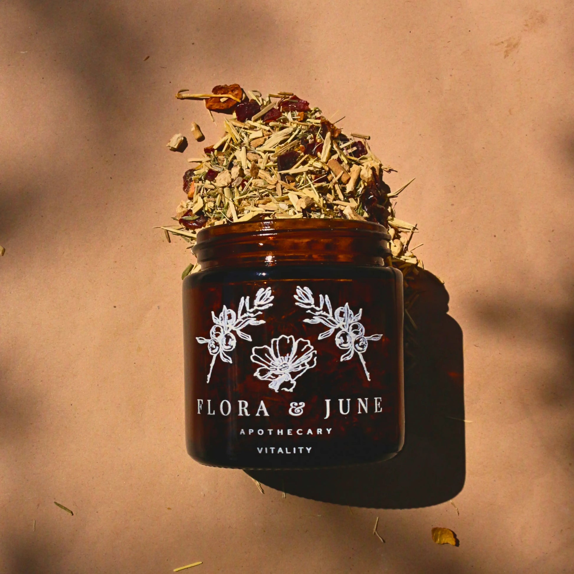 An amber glass jar on it's side with Flora & June's Vitality loose leaf tea spilling out onto the page. The jar has a white label showing the brand name "Flora & June Apothecary" and the brand logo of a blooming flower and two sprigs of Juniper branch.